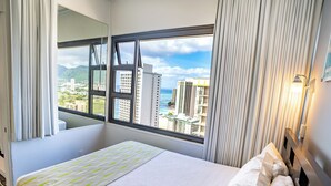 A hotel room with a large window, a bed with white bedding, and a view of buildings and mountains.