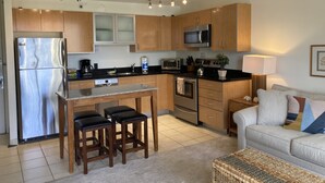A modern kitchen with a stainless steel refrigerator, microwave, and oven. There is a bar area with stools, a coffee machine, and a sink. A sofa and a lamp are in the living area. - Deluxe Condo , Free Parking & Wi-Fi (Honolulu)