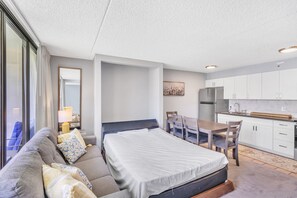 Quarto compacto com cama, sofá, mesa de jantar com cadeiras, cozinha e uma janela grande com cortinas.