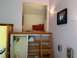 A room with a loft bed, wooden ladder, and a wall-mounted shelf.