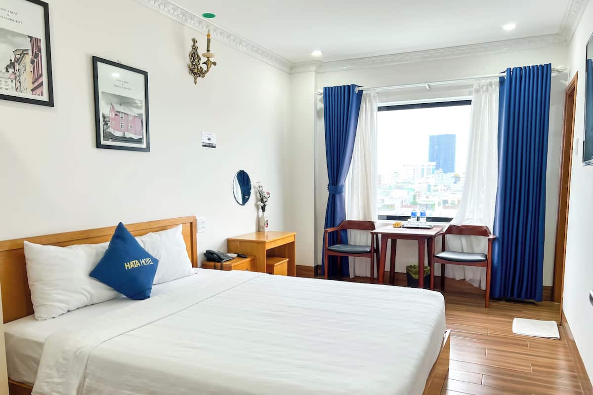 A hotel room with a large bed, a desk with a chair, a window with curtains, and framed pictures on the wall.