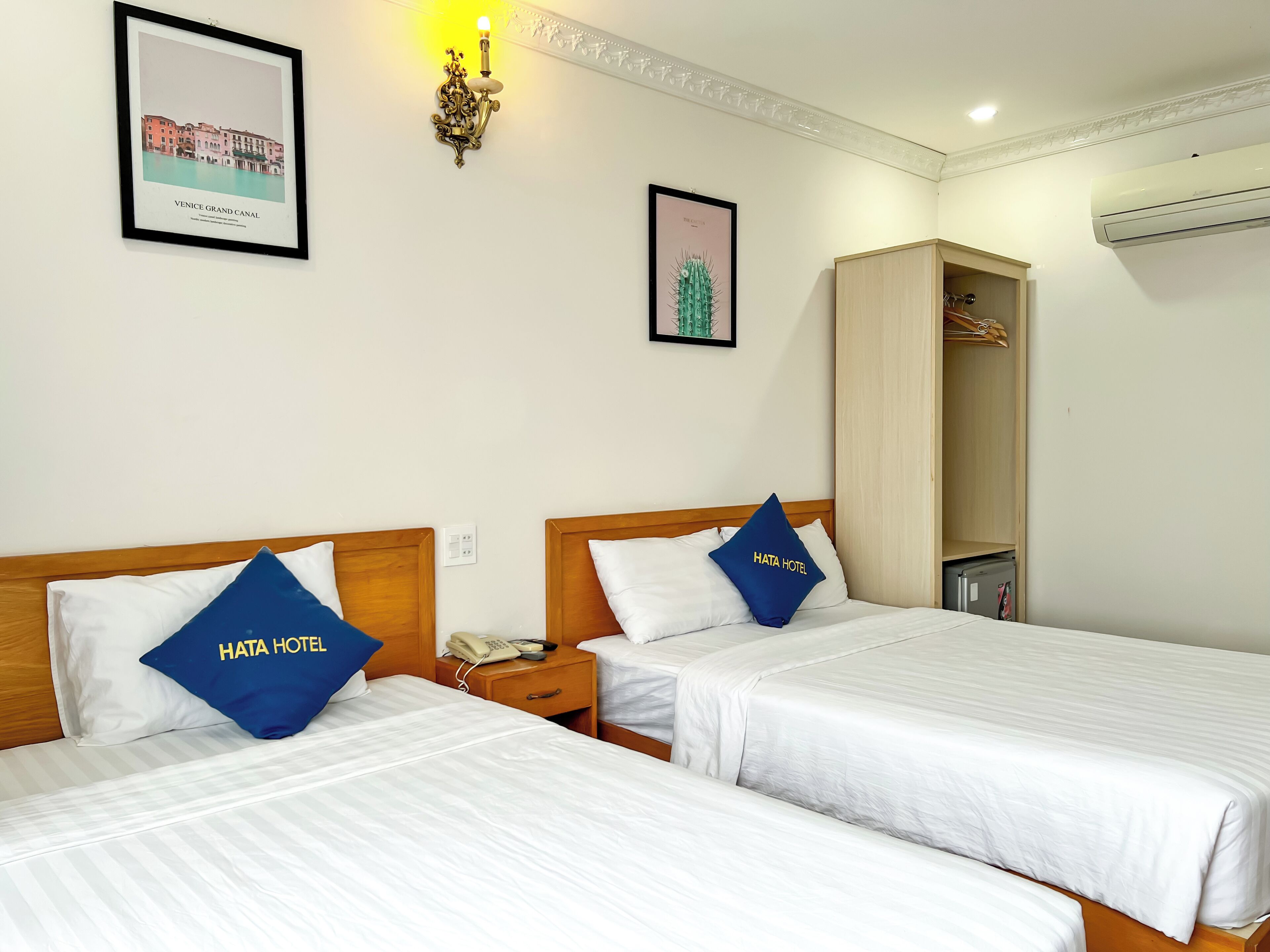 A hotel room with two beds, each with a pillow labeled "HATA HOTEL". There is a framed picture of the Venice Grand Canal on the wall, a wall-mounted lamp, and an air conditioning unit.