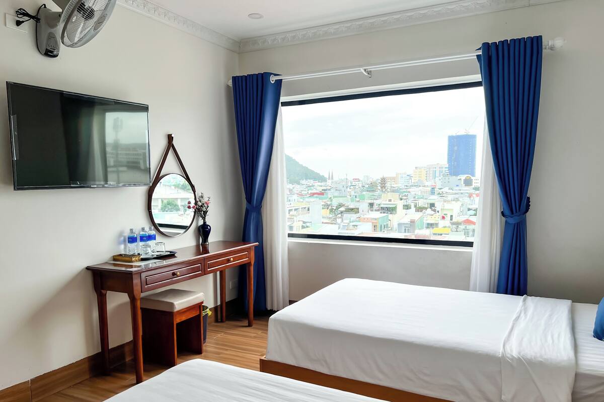 A hotel room with two beds, a desk with a mirror, a flat-screen TV, and a window with blue curtains.