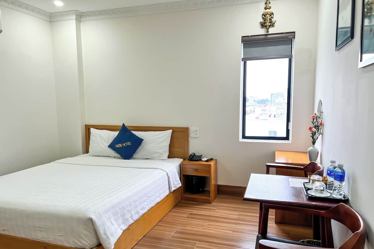 A hotel room with a bed, a desk, a chair, a window with a city view, and a vase with flowers.