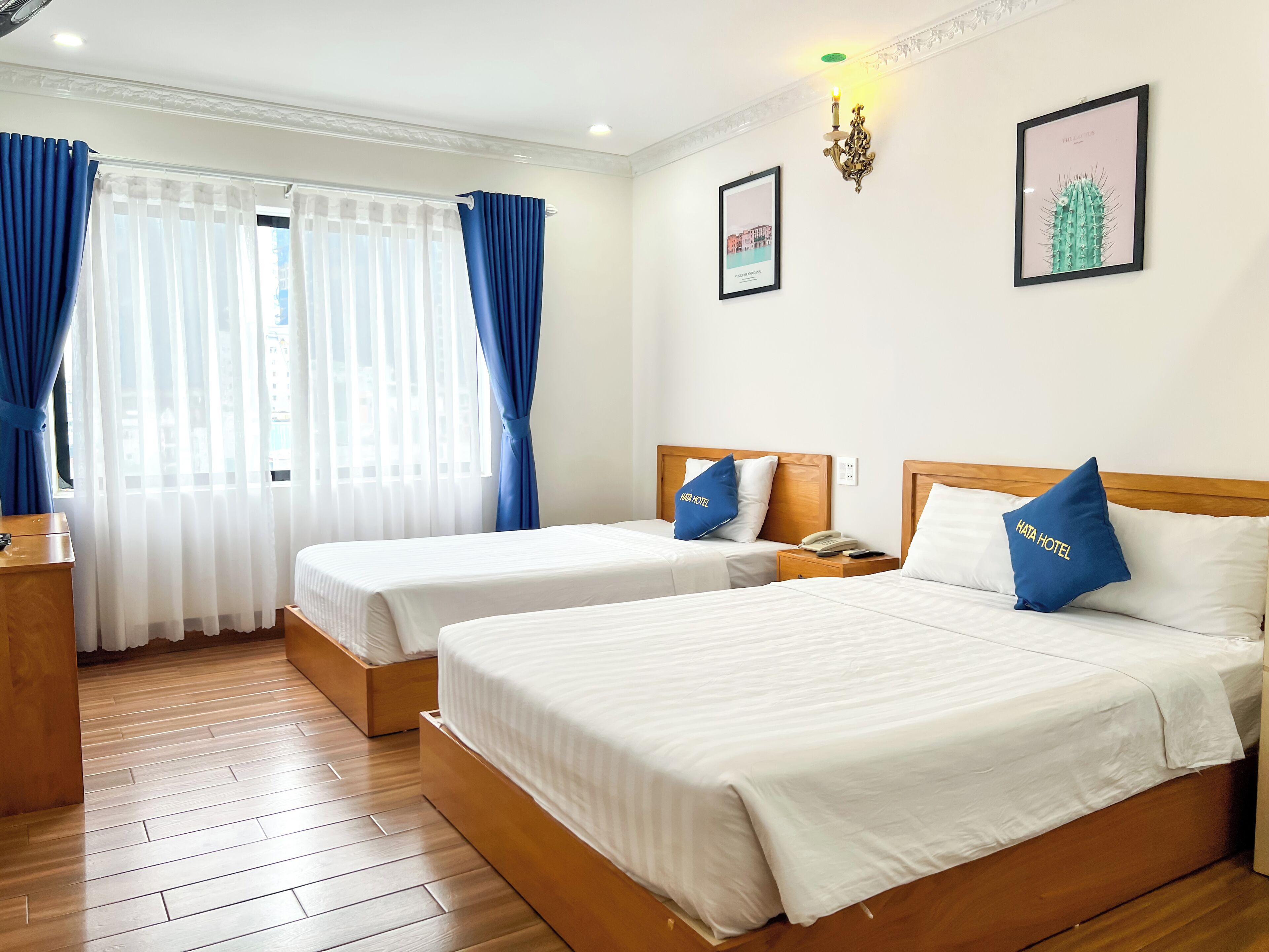 A hotel room with two beds, a wooden floor, a window with blue curtains, a wall-mounted phone, and framed pictures.