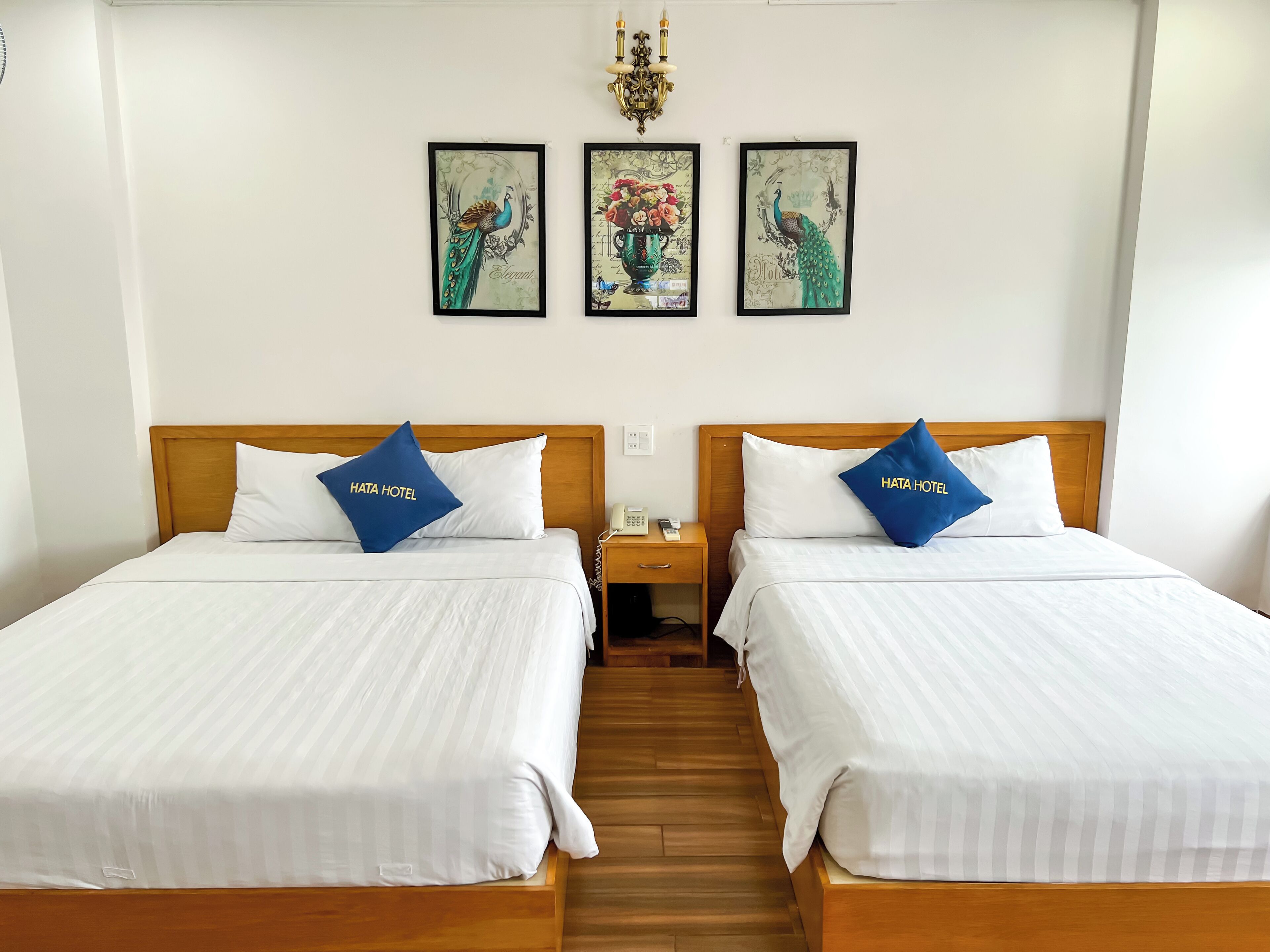 A hotel room with two beds, each with a pillow labeled "HATA HOTEL," a wooden headboard, a nightstand with a phone, and a wall with three framed pictures.