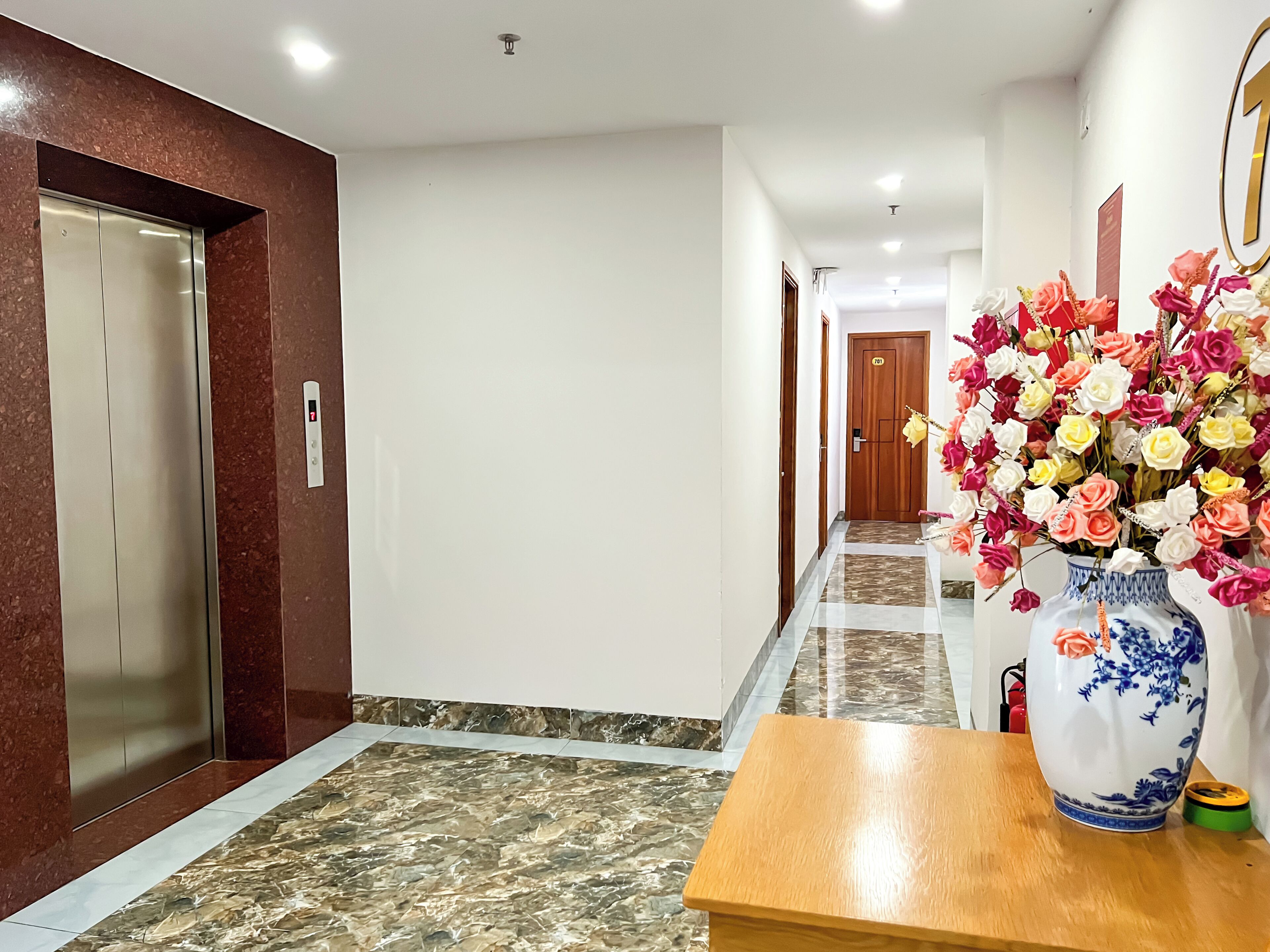 A hotel hallway with a marble floor, an elevator, a wooden door, and a vase of flowers on a table.