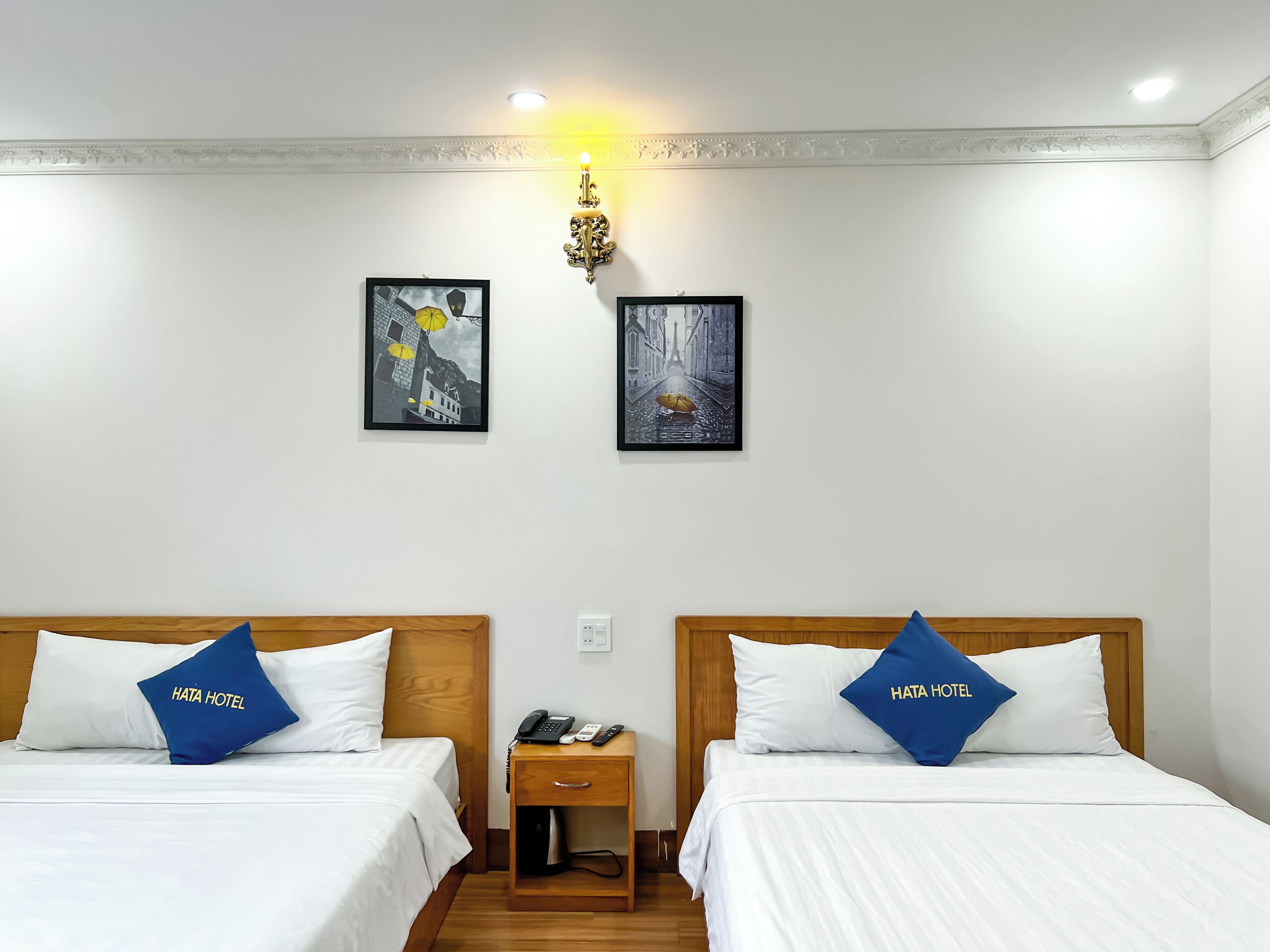 A hotel room with two beds, each with a pillow labeled "HATA HOTEL," a bedside table with a phone and a lamp, and two framed pictures on the wall.