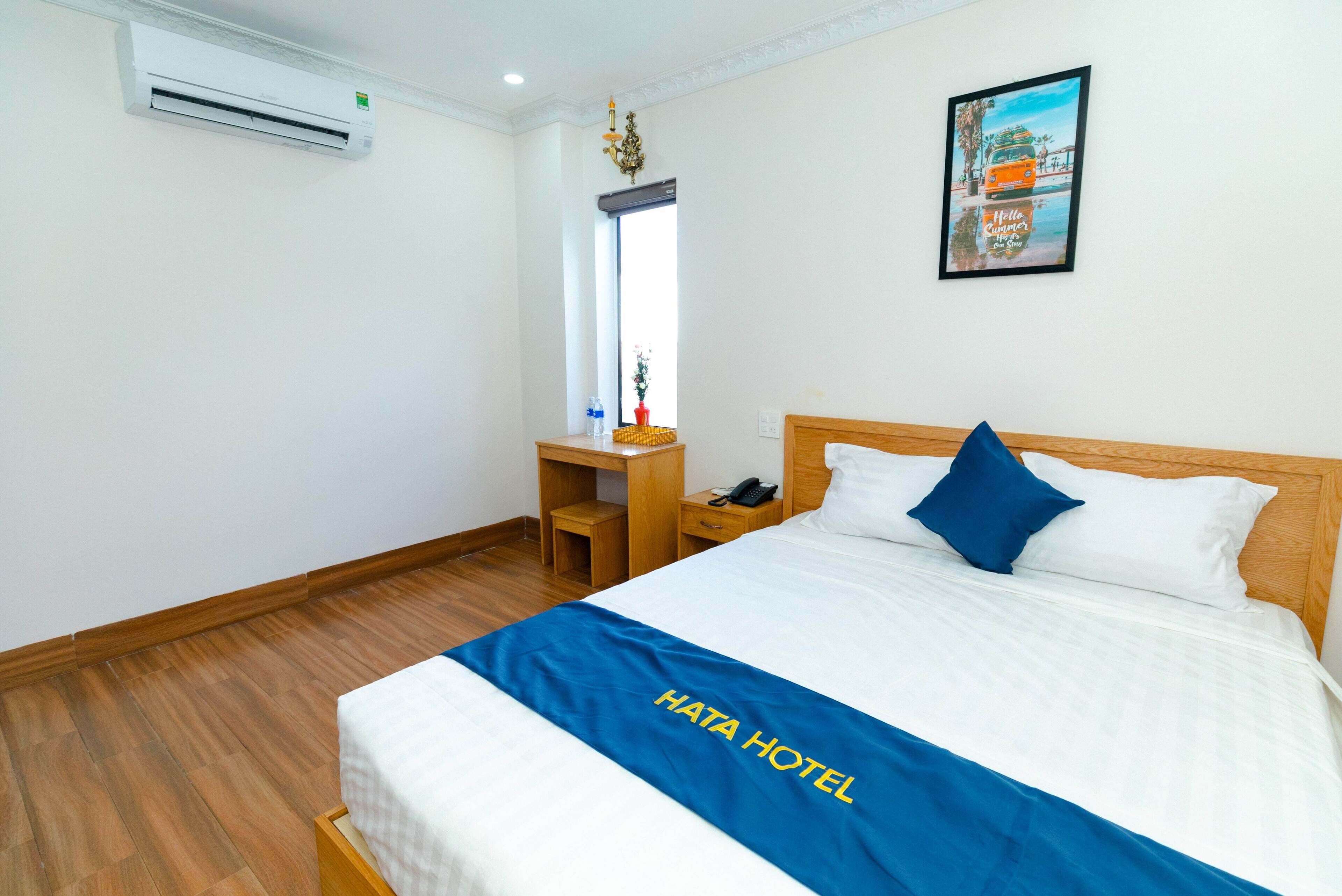 A hotel room with a bed, a wooden nightstand, a desk, an air conditioner, a window, and a framed picture on the wall.