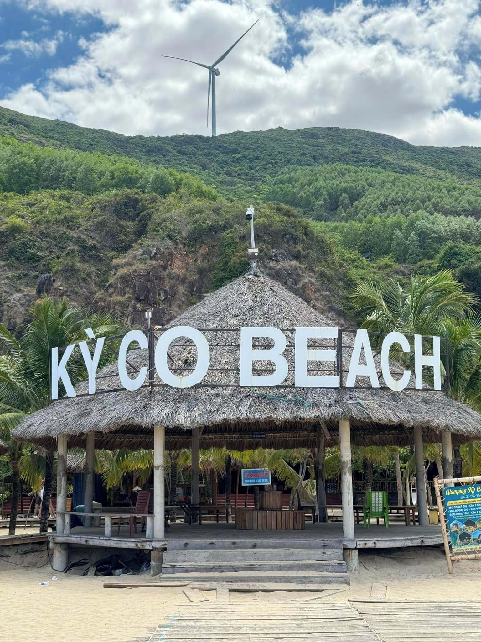 Un abri de plage avec une pancarte indiquant « KYCO BEACH » et une éolienne en arrière-plan.