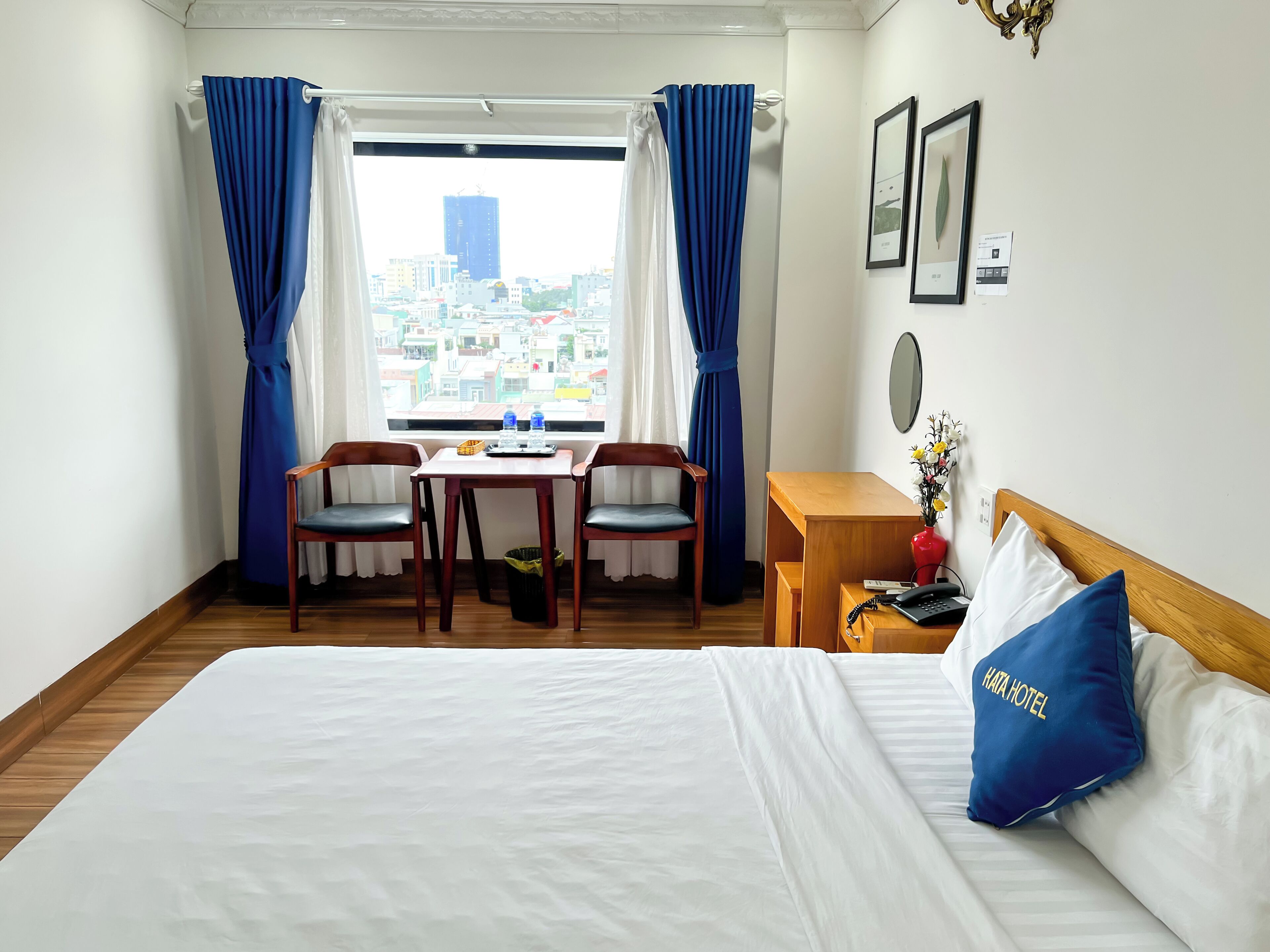 A hotel room with a bed, a desk with two chairs, a view of the city, and a window with blue curtains.