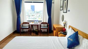 Habitación de hotel con cama, escritorio y dos sillas, vista a la ciudad y ventana con cortinas azules.