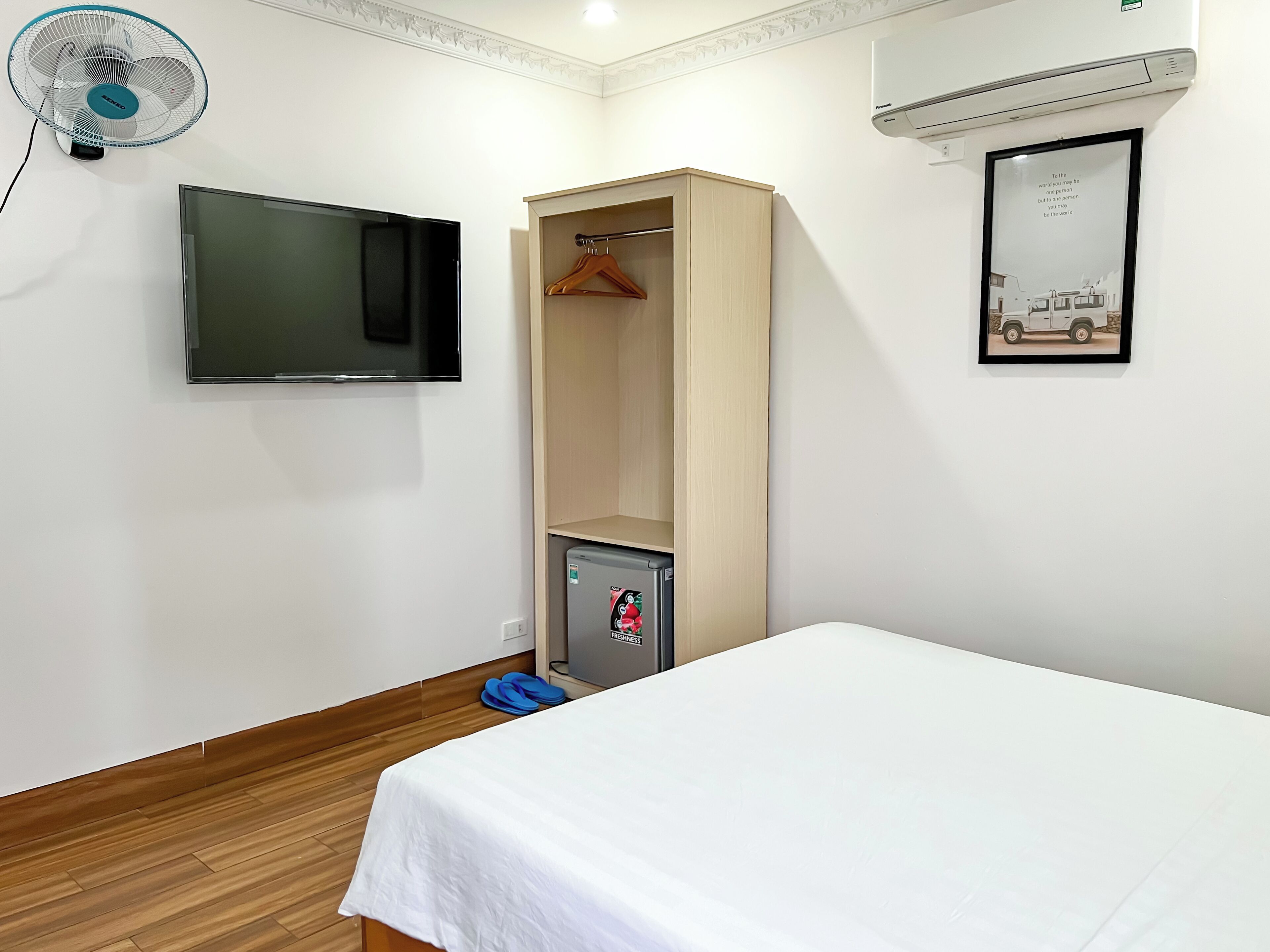A bedroom with a bed, a TV, a wardrobe, a small fridge, and a wall-mounted air conditioner.