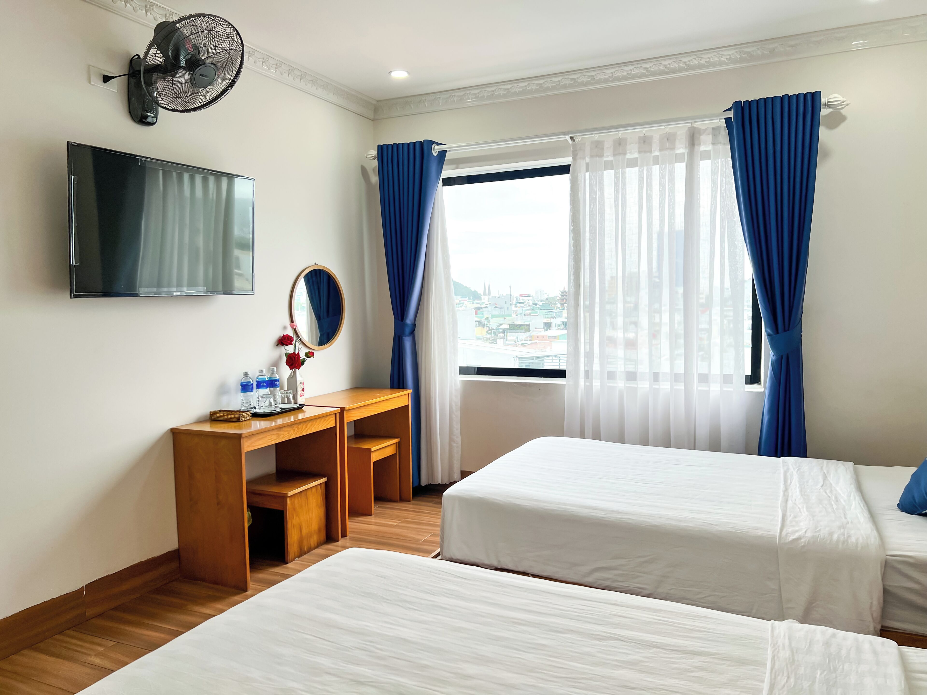 A hotel room with two beds, a desk with a mirror, a TV, and a window with blue curtains.