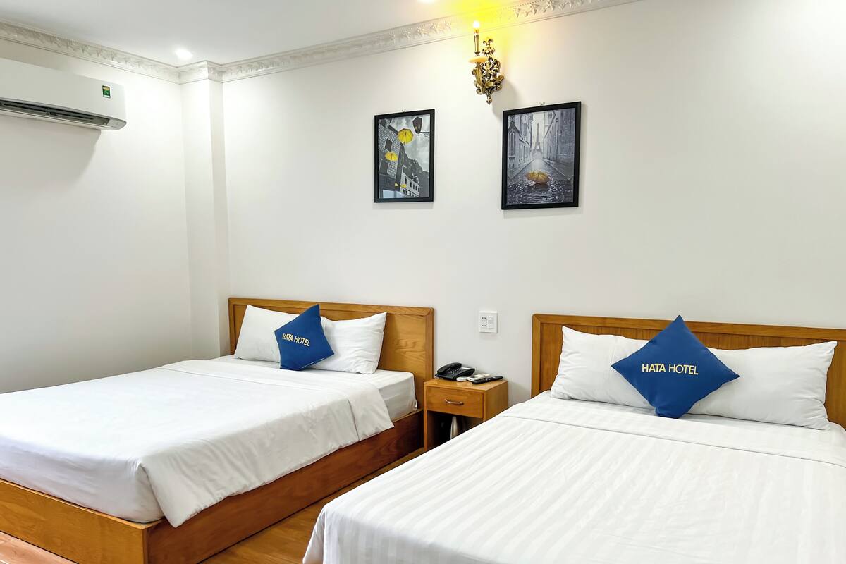 A hotel room with two beds, each with a wooden headboard and white bedding. There are two framed pictures on the wall, a nightstand with a phone and remote, and an air conditioning unit on the wall.