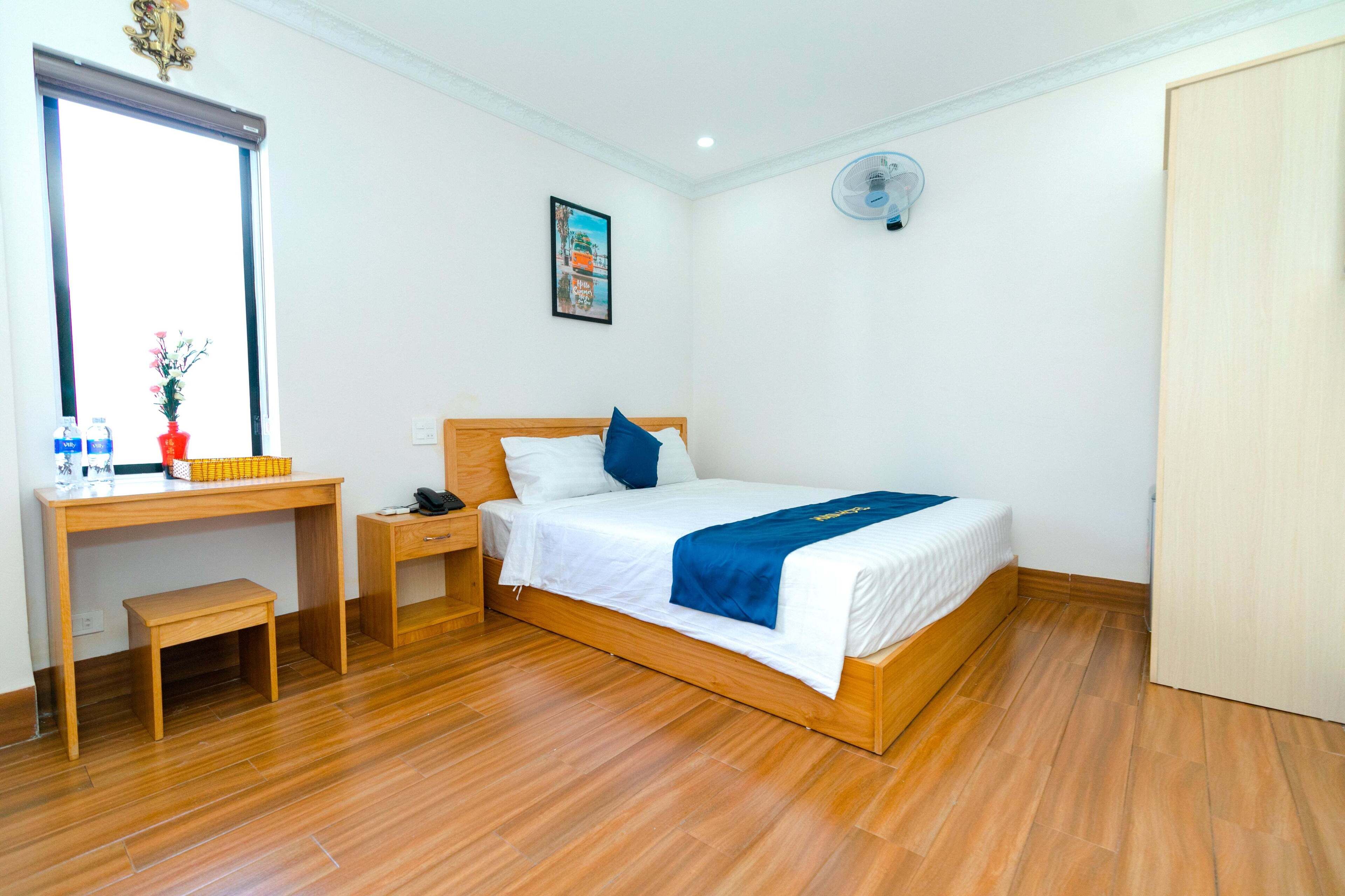 A hotel room with a wooden bed, a desk, a chair, a small table, a nightstand, and a window with a view.