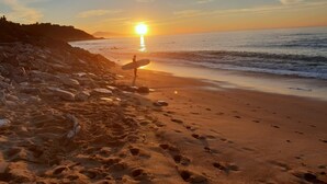 Eine Strandansicht mit Fußspuren, die zum Wasser führen, einer Person, die einen Surfbretträger hält, und einer untergehenden Sonne über dem Meer.