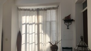 Ein Zimmer mit großem Fenster, das mit durchsichtigen Vorhängen versehen ist, einer Topfpflanze, einem Sessel, einem kleinen Tisch und einem wandmontierten Regal mit einem dekorativen Gegenstand.