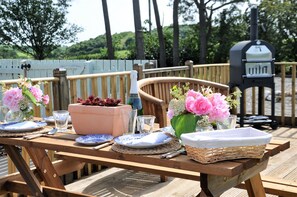 A wooden deck with a table set for dining, featuring blue and white plates, clear glasses, a bottle, and a vase with pink flowers.