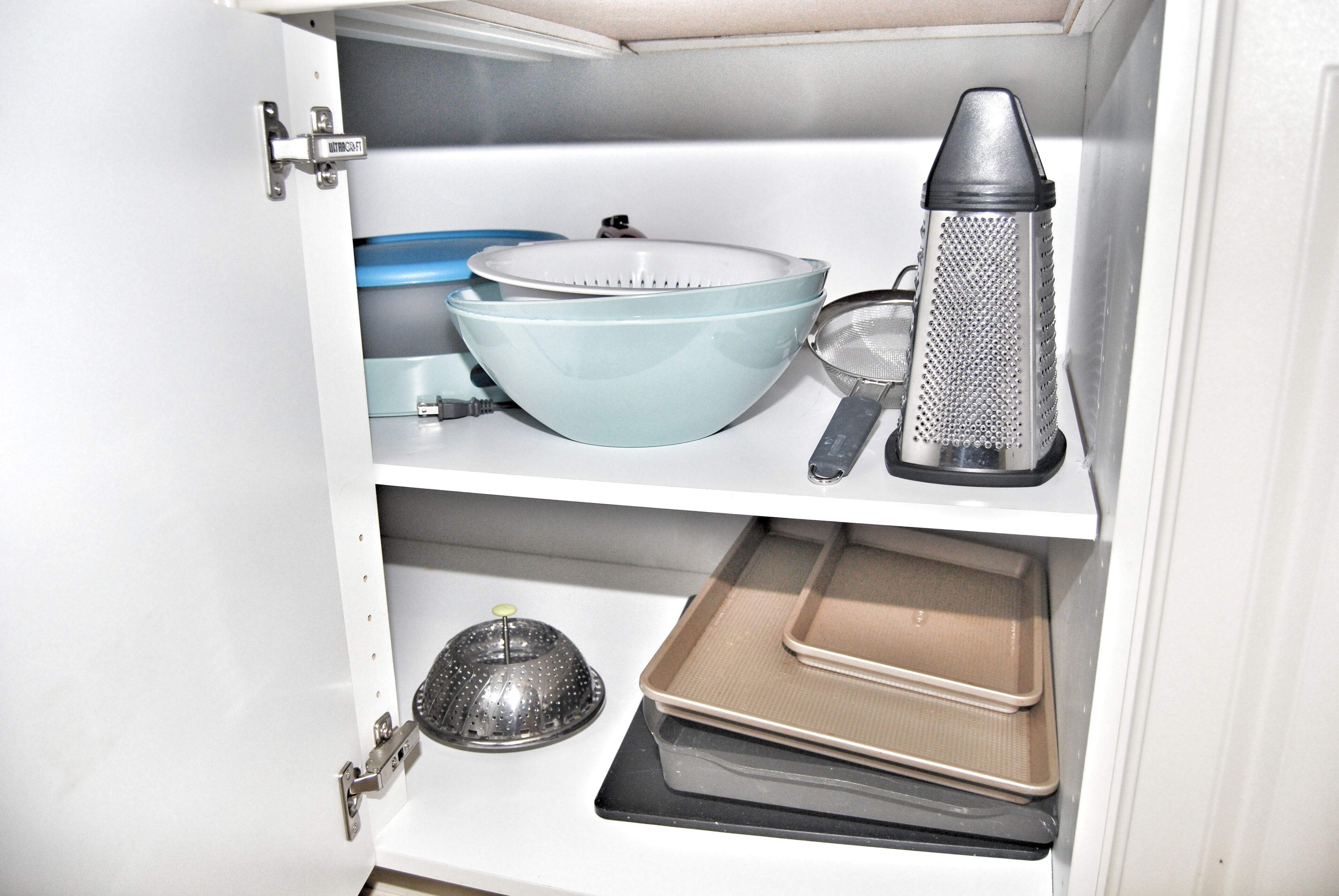 A kitchen cabinet with various utensils and cookware, including a grater, a strainer, and baking trays.