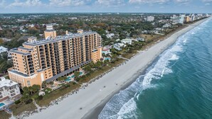 An aerial view of a coastal hotel with multiple floors, a beach adjacent to the property, and a clear view of the ocean.