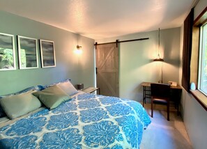 Une chambre à coucher avec un lit, un bureau et une chaise. Il y a trois lampes fixées au mur, une fenêtre avec des rideaux et une porte également dotée d’un rideau.