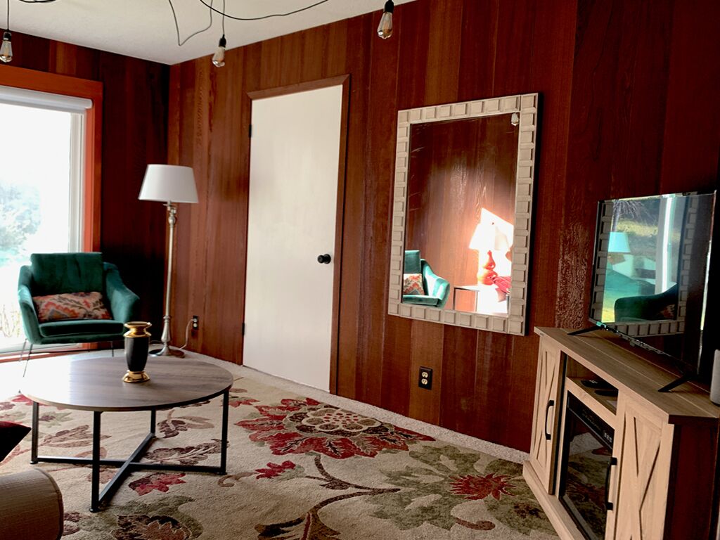 Un salón con un sillón verde, una mesa redonda de centro, una alfombra estampada, un televisor sobre un mueble de madera y un espejo en la pared.