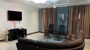 Una sala de estar moderna con un sofá de cuero negro, dos sillones negros, un televisor de pantalla plana, un ventilador de techo y un ventanal con cortinas.