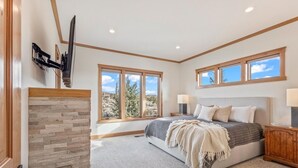 Un dormitorio con chimenea de piedra, cama con ropa de cama gris y blanca, dos ventanas y cabecera de madera.