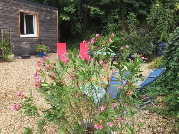 Une cabane en bois avec une fenêtre, entourée d’un jardin où poussent des fleurs roses et où se trouve une chaise bleue.