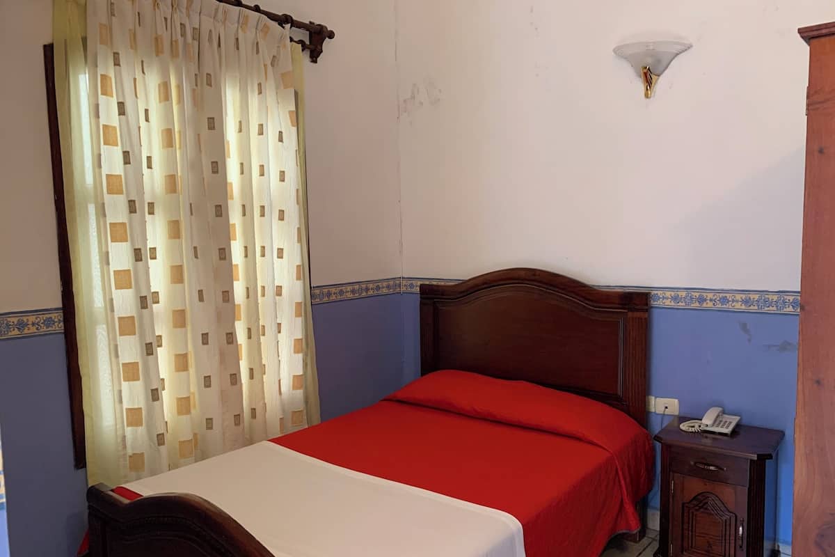 Ein Schlafzimmer mit einem hölzernen Bett, einer roten Bettdecke, einer weißen Decke, einem Nachttisch mit einem Telefon und einer wandmontierten Leuchte.