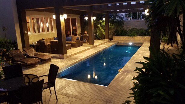 Una piscina con sillas de descanso y mesas, rodeada de plantas y un área de estar techada con luces.