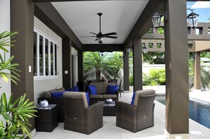 Relax in a shaded seating area with wicker chairs and a sofa, overlooking a tranquil pool.