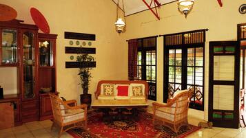 A traditional room with a sofa, two wicker chairs, a red patterned rug, a wooden cabinet, and a potted plant.