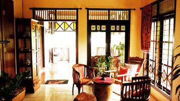 A room with a tiled floor, wooden furniture, a large plant, and a view through a window with a patterned curtain.