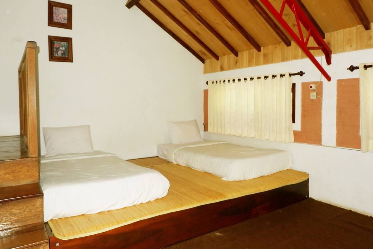 A room with two single beds, wooden flooring, a sloped ceiling with exposed beams, and a window with curtains.