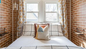 Une chambre avec un mur en briques, deux fenêtres et un lit avec des oreillers.