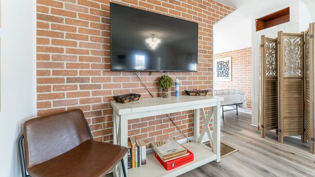 Un salon avec un mur en briques, un téléviseur à écran plat fixé au-dessus d’une console blanche, un fauteuil en cuir marron et un canapé blanc.