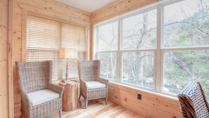 A cozy room with wooden walls and large windows offering a view of trees. There are two wicker chairs with cushions.