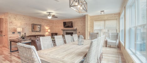 Un comedor espacioso con una larga mesa de madera, sillas de mimbre, una chimenea y un amplio ventanal con persianas.