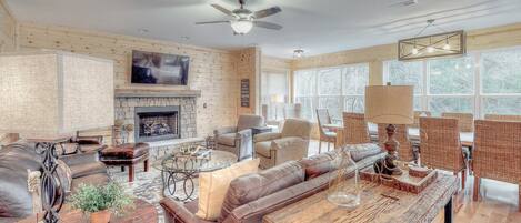 A spacious living room with a fireplace, leather furniture, wicker chairs, and a large window with a view of trees.
