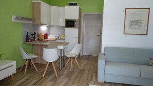 Un ambiente compatto con angolo cottura, zona pranzo con due sedie, divano e un quadro appeso al muro.