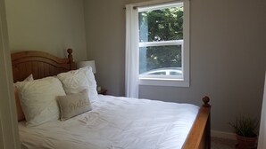 Une chambre à coucher avec une tête de lit en bois, du linge de lit blanc, une fenêtre donnant sur des arbres et une plante en pot.
