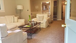 Un salon avec un canapé blanc, une table basse et une lampe. Une porte donne accès à une autre pièce.