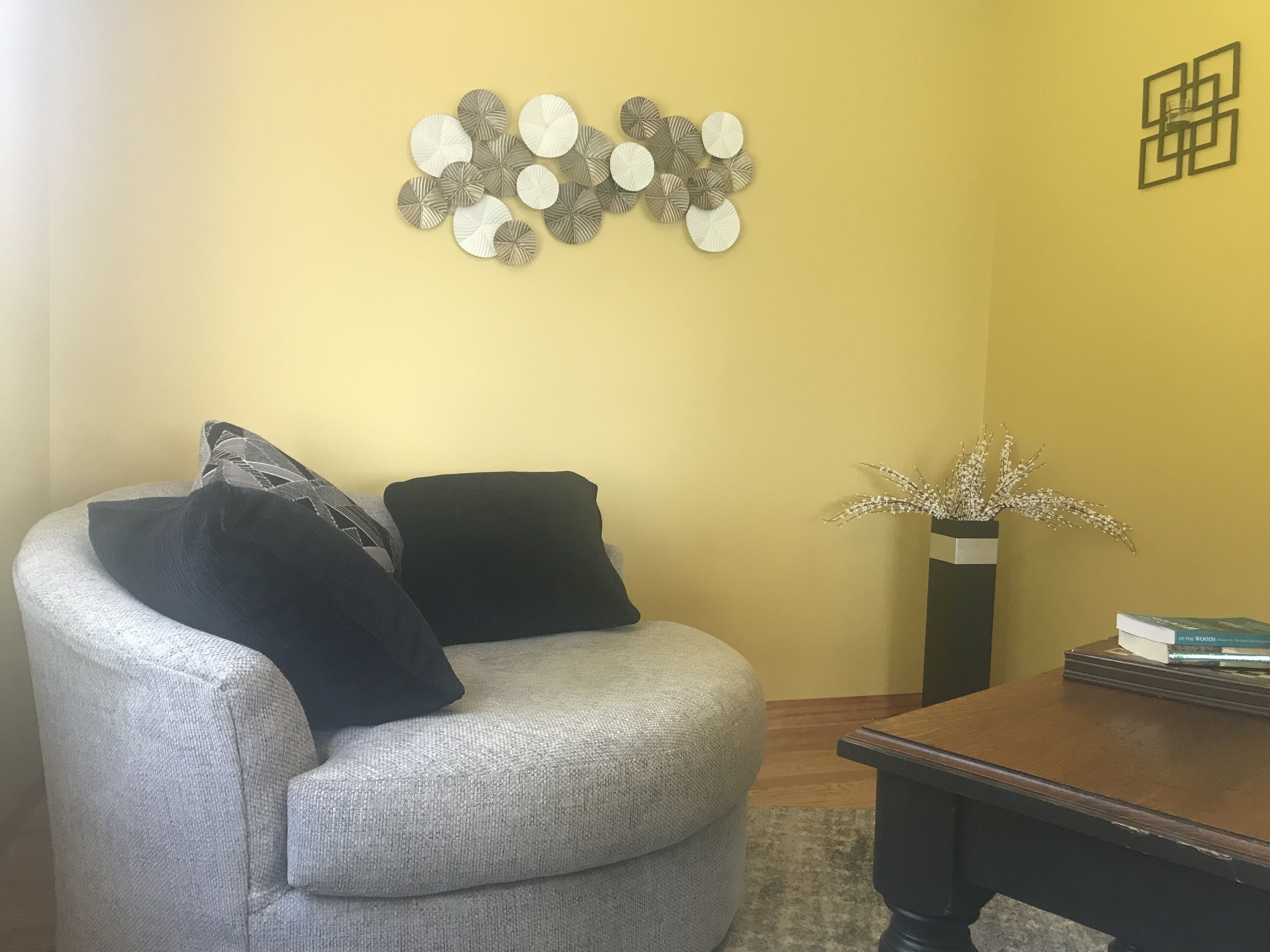 A living room with a gray sofa, two black pillows, a wooden coffee table, a vase with white flowers, and a wall art piece.