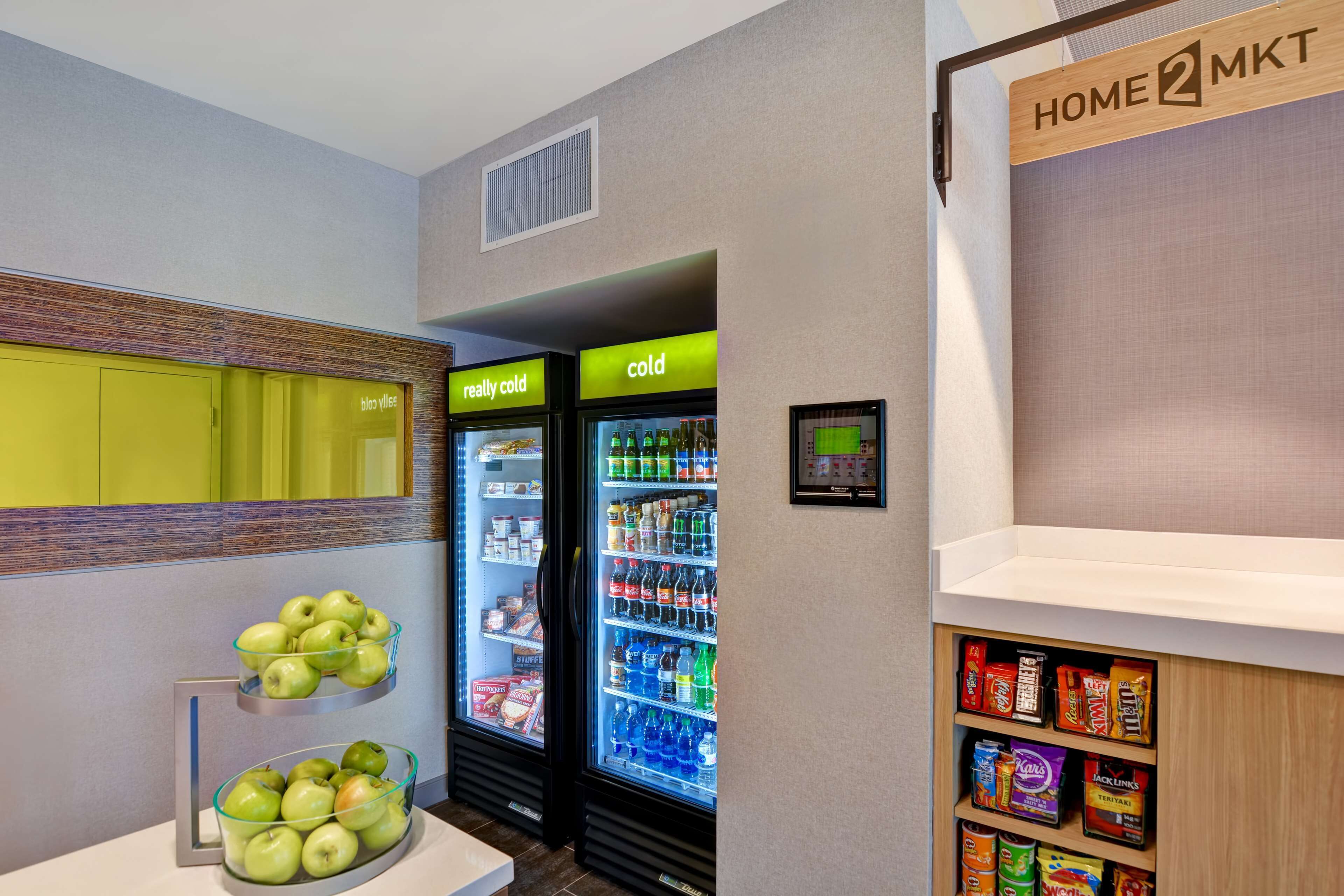Eine Hotelküche mit Obstschale, zwei Getränkespendern, auf denen 'wirklich kalt' und 'kalt' steht, und einem Regal voller Snacks.