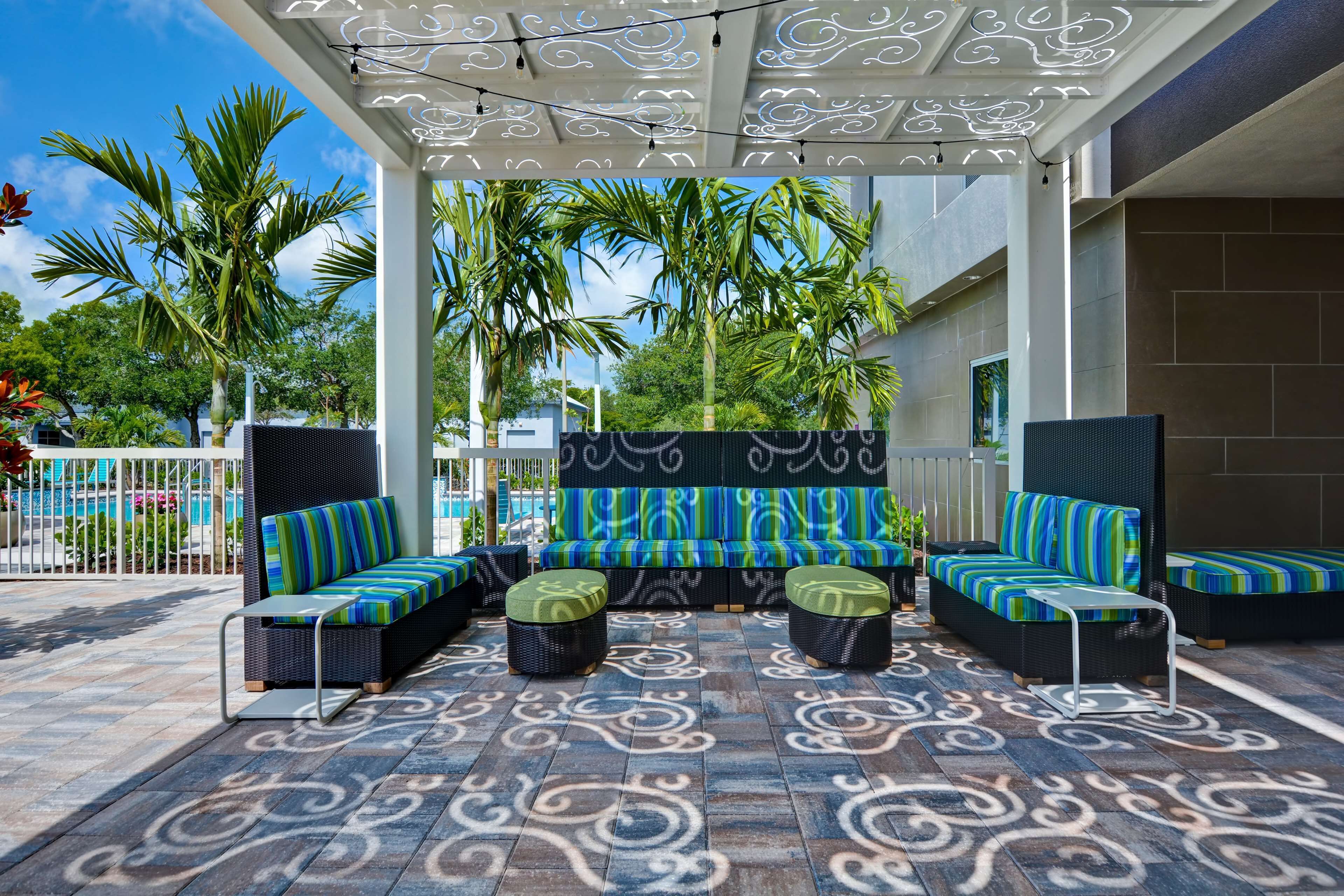 A covered outdoor seating area with patterned flooring, wicker furniture, and a view of palm trees and a pool.