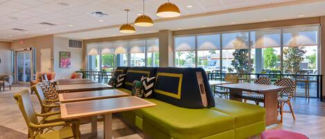 A spacious indoor area with a green sofa, wooden tables, wicker chairs, and large windows offering a view of the outside.