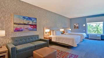 A hotel room with a sofa, two beds, a nightstand, a lamp, a painting, and a window with a view of palm trees.