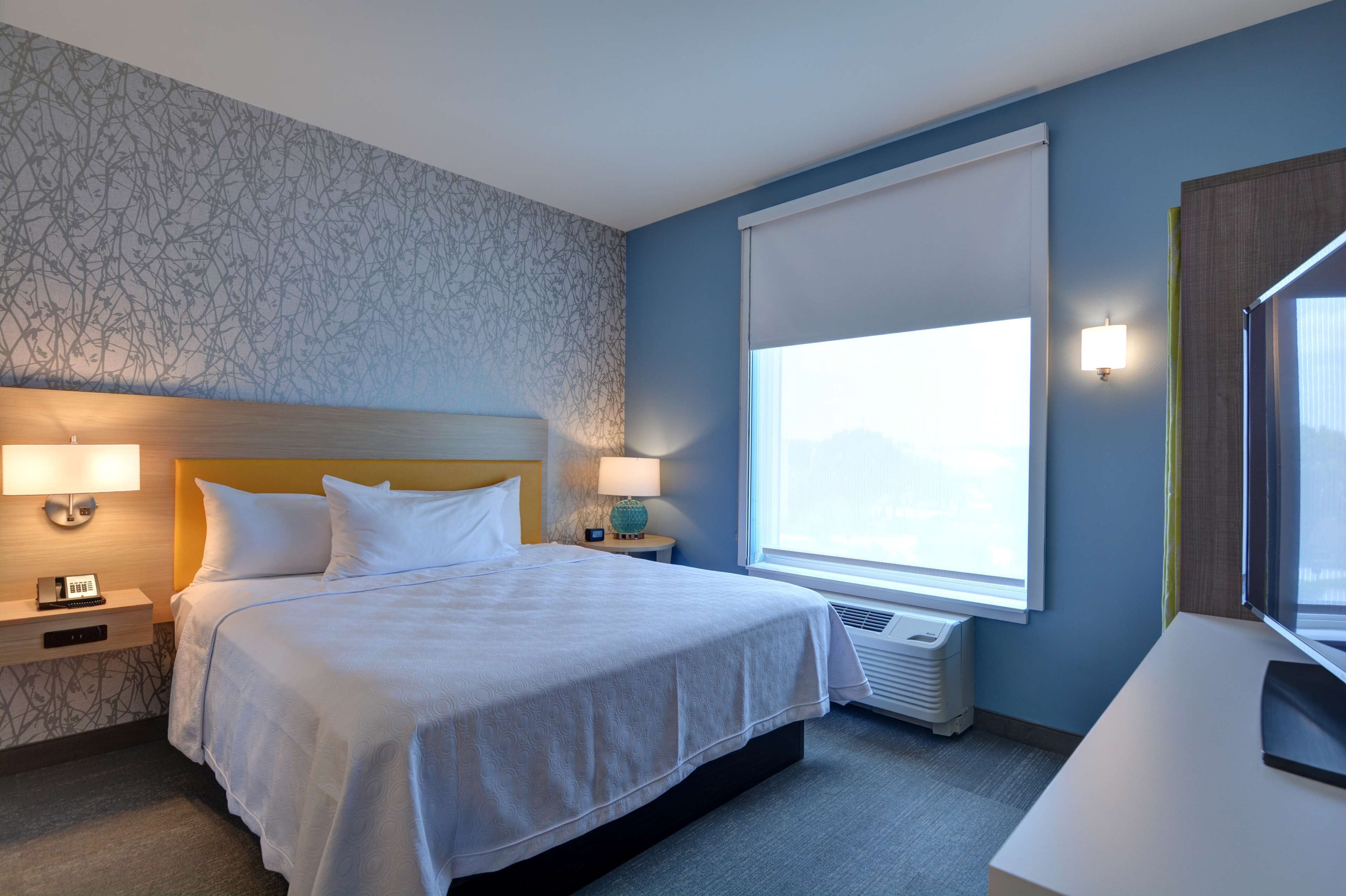 Une chambre d’hôtel avec un grand lit, deux tables de chevet avec des lampes, une fenêtre avec des stores et un bureau avec une chaise.